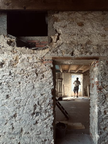 Intérieur en rénovation avec murs en pierre apparente, poutre en bois et ouvertures traversantes vers l'extérieur