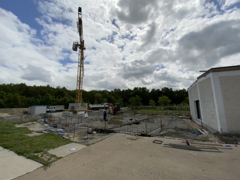 Chantier de construction avec fondations en béton armé, grue, pelleteuse et bâtiment existant en briques blanches