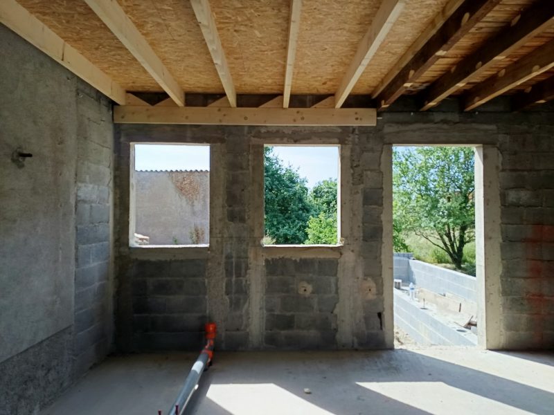Espace intérieur en construction avec murs en béton brut, charpente bois apparente et ouvertures donnant sur jardin verdoyant