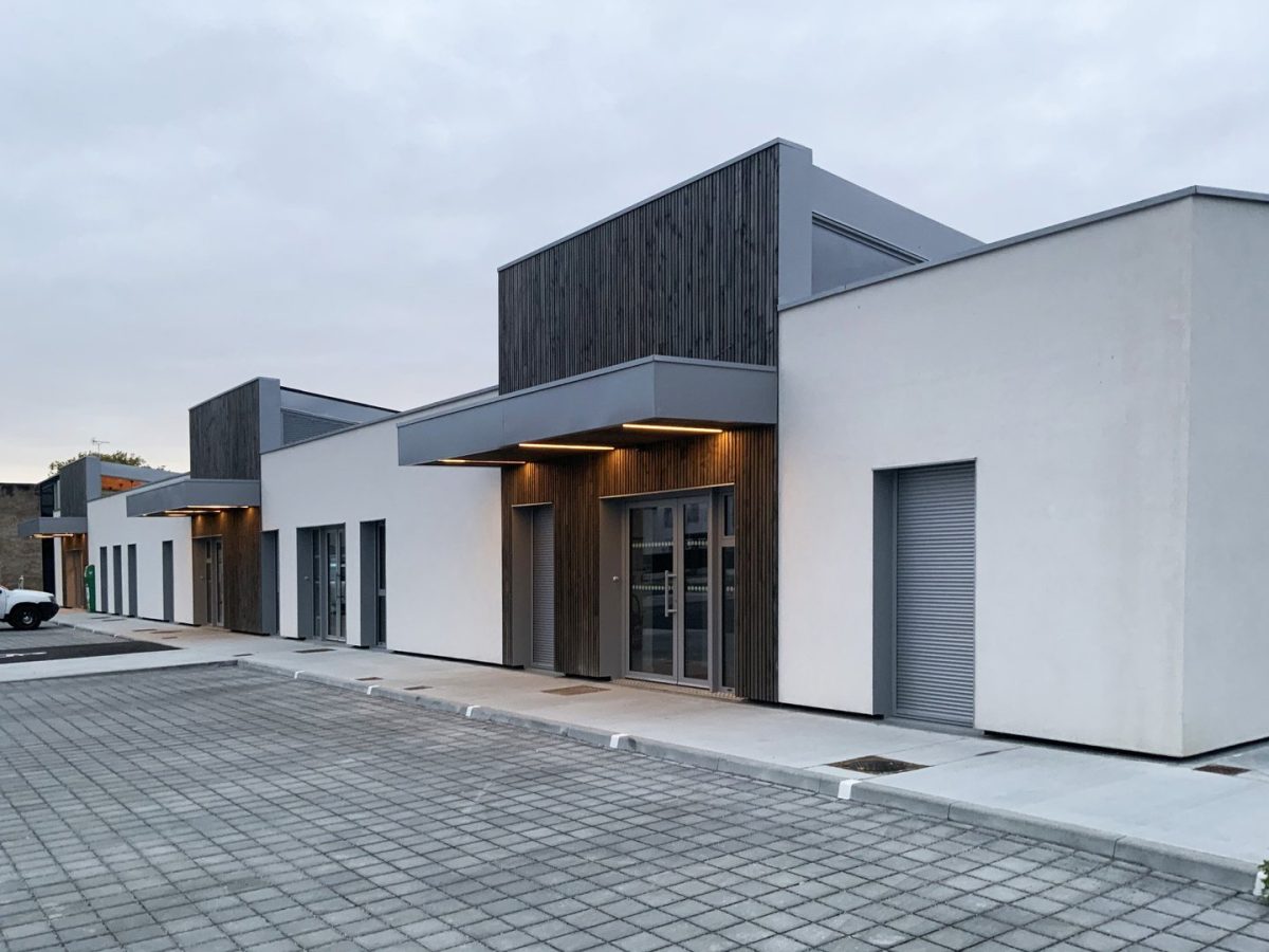 Ensemble de bâtiments modernes avec bardage bois vertical, façades blanches et volets roulants sur parking pavé