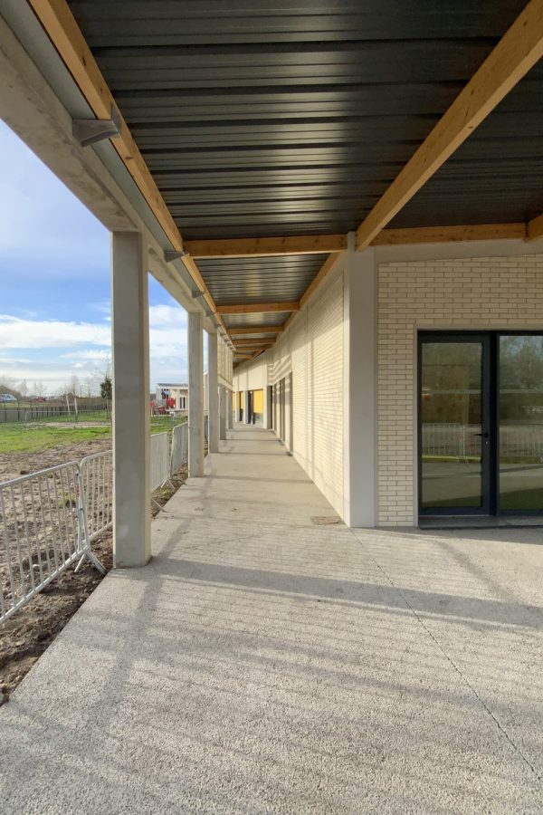 Coursive couverte avec poutres bois et plafond métallique, murs en briques blanches, sol béton, rambarde métallique