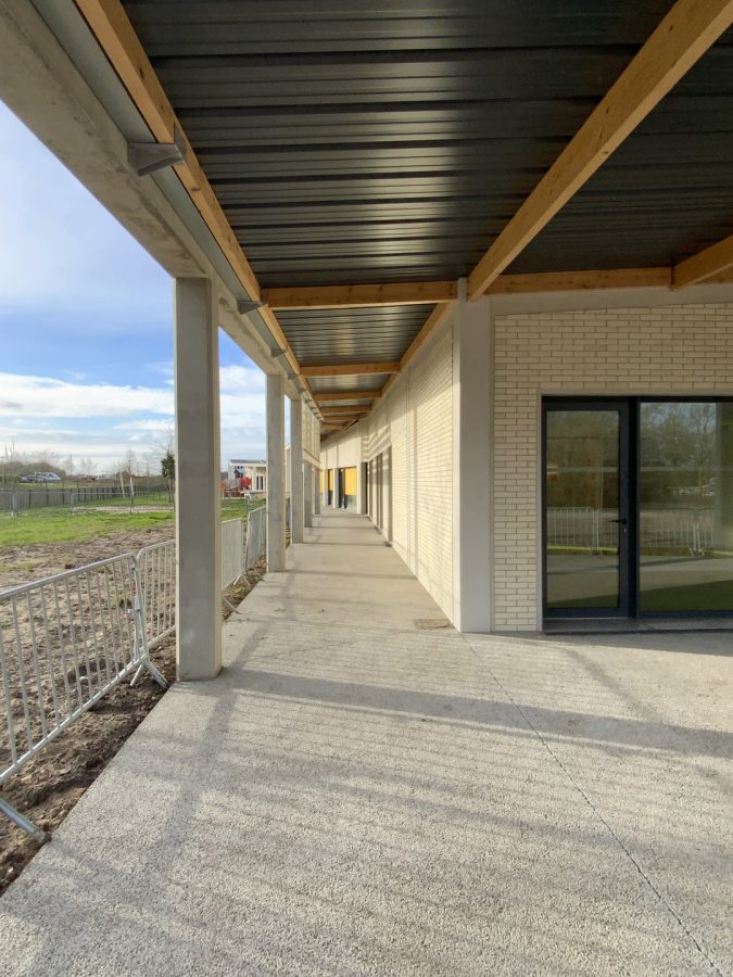 Coursive couverte avec poutres bois et plafond métallique, murs en briques blanches, sol béton, rambarde métallique