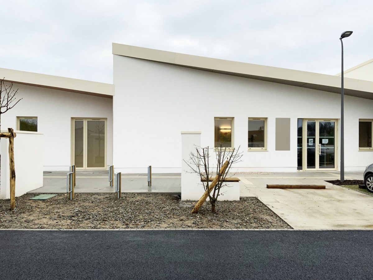 Bâtiment moderne blanc à toit plat avec grandes fenêtres, cour en gravier avec plantations et éclairage urbain