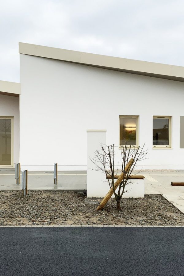 Bâtiment moderne blanc à toit plat avec grandes fenêtres, cour en gravier avec plantations et éclairage urbain