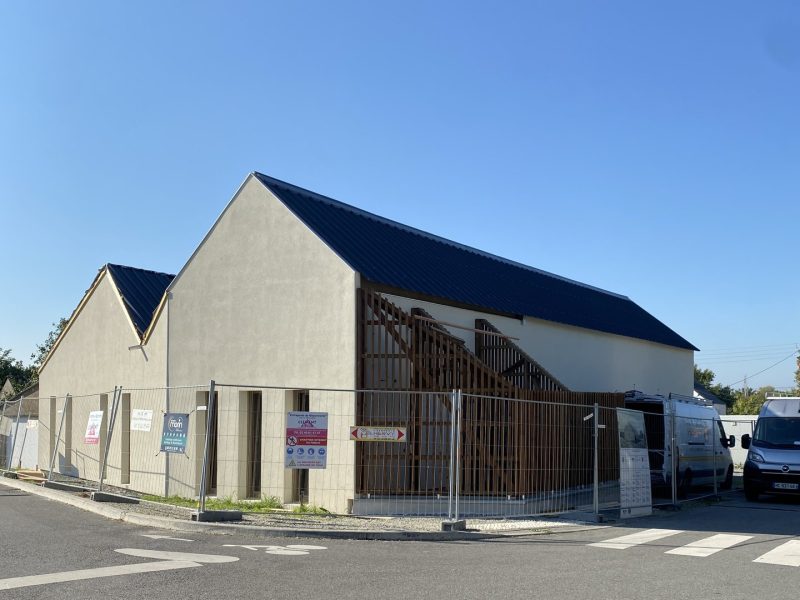 Bâtiment commercial neuf avec façade enduite beige et toiture anthracite, entouré de clôtures de chantier temporaires