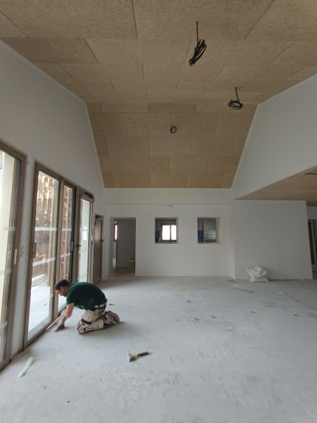 Grande pièce en construction avec plafond en panneaux de bois clair, murs blancs et ouvrier au sol près de baies vitrées