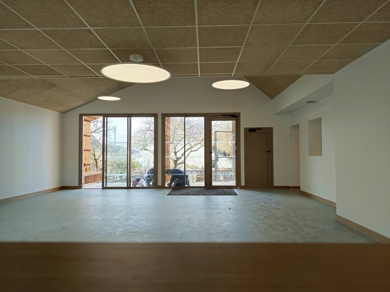 Salle vide avec plafond acoustique, sol béton poli et grandes baies vitrées donnant sur un arbre et une terrasse extérieure