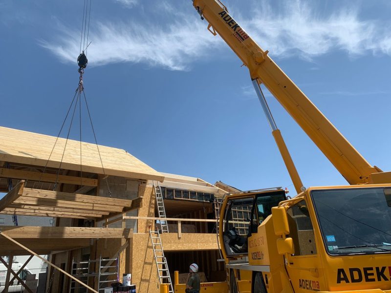 Chantier de construction en bois avec grue jaune soulevant une poutre, structure à ossature bois et panneaux OSB