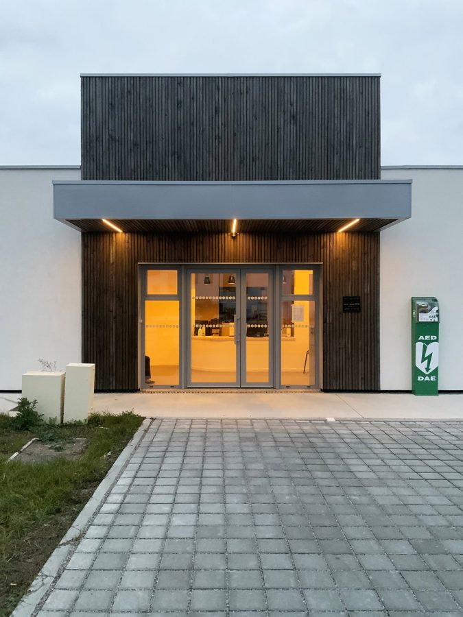 Entrée moderne d'un bâtiment avec bardage bois foncé, porte vitrée illuminée et defibrillateur extérieur