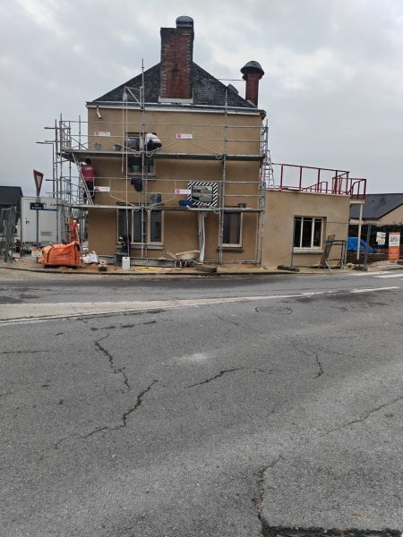 Maison en rénovation avec échafaudage, enduit beige, toit en ardoise et cheminées en brique, au coin d'une rue