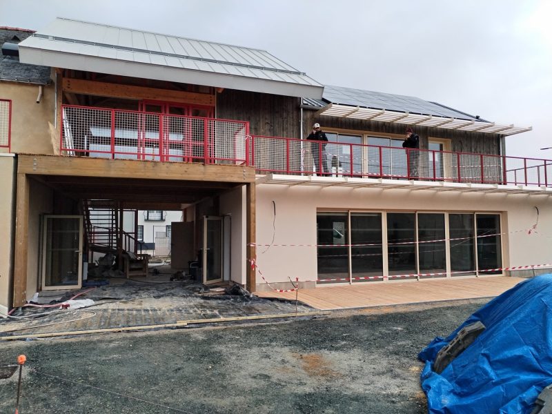 Maison contemporaine en construction avec bardage bois, balcon à garde-corps rouge et panneaux solaires en toiture