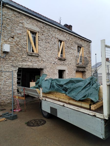 Maison en pierre ancienne en rénovation avec fenêtres condamnées, camionnette de chantier chargée de matériaux au premier plan