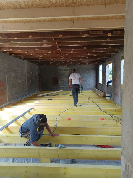 Chantier de rénovation avec isolation jaune au sol, poutres apparentes et murs en parpaings. Deux ouvriers travaillent.