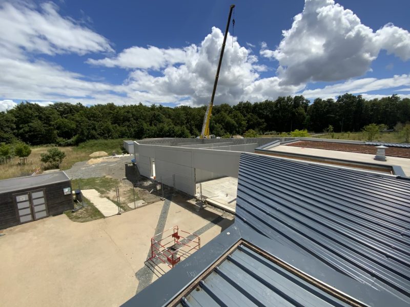 Vue aérienne d'un chantier de construction avec grue, bâtiments modernes à toiture métallique et environnement boisé