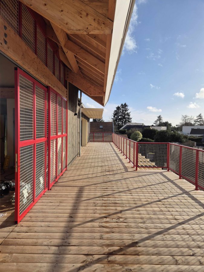 Terrasse en bois avec garde-corps rouge en métal et treillis, structure en bois apparente, vue sur quartier résidentiel