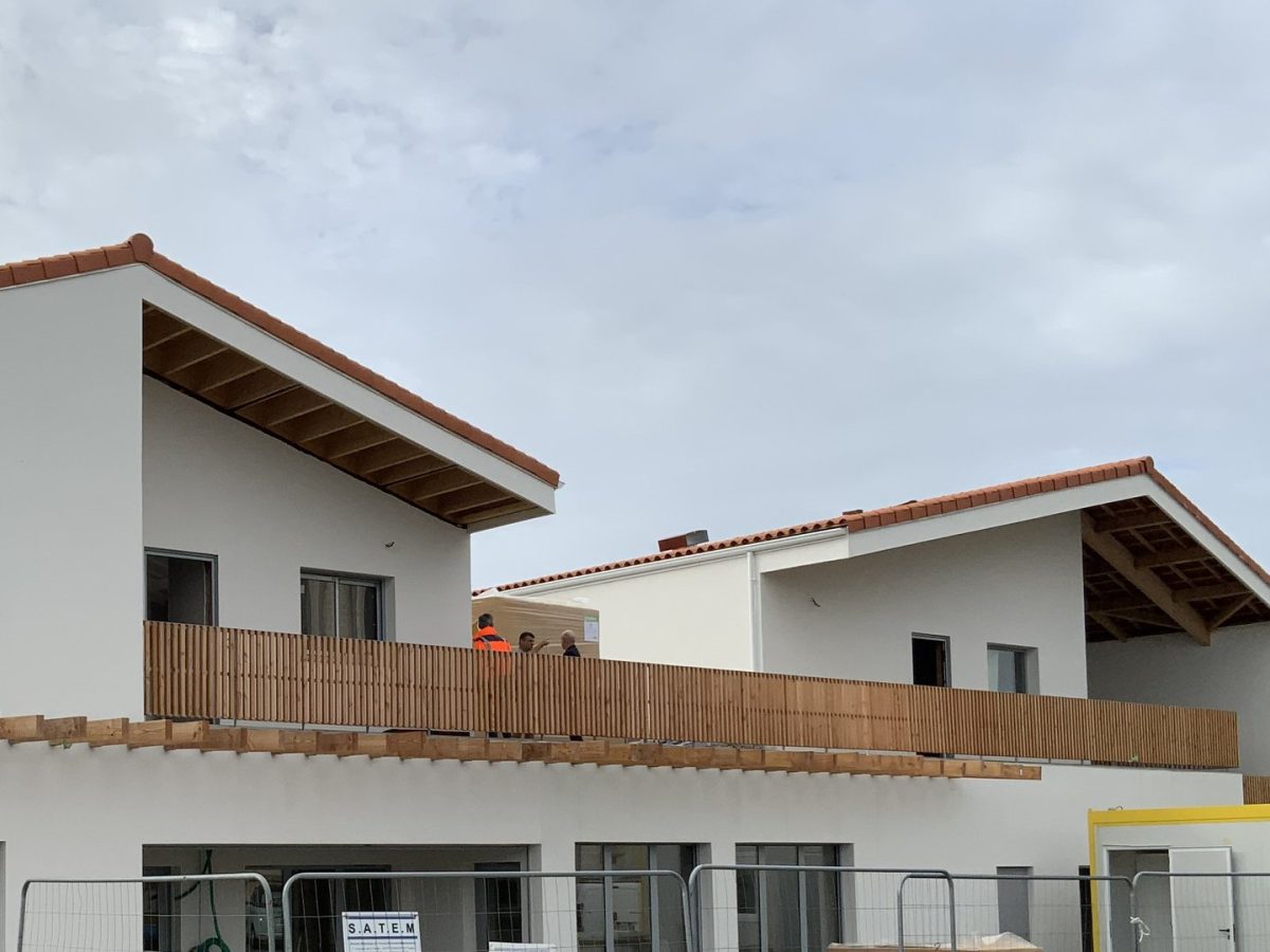 Bâtiment moderne à toiture tuiles terre cuite, façades blanches et garde-corps bois, en construction avec barrières chantier