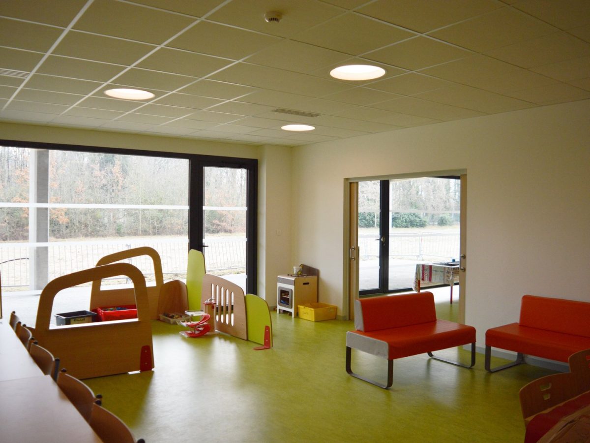 Salle de jeux lumineuse avec sol vert, mobilier coloré, plafond acoustique et grandes baies vitrées donnant sur l'extérieur