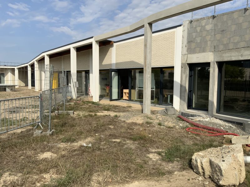 Bâtiment en construction avec façade en béton, briques beiges et grandes baies vitrées, terrain en travaux