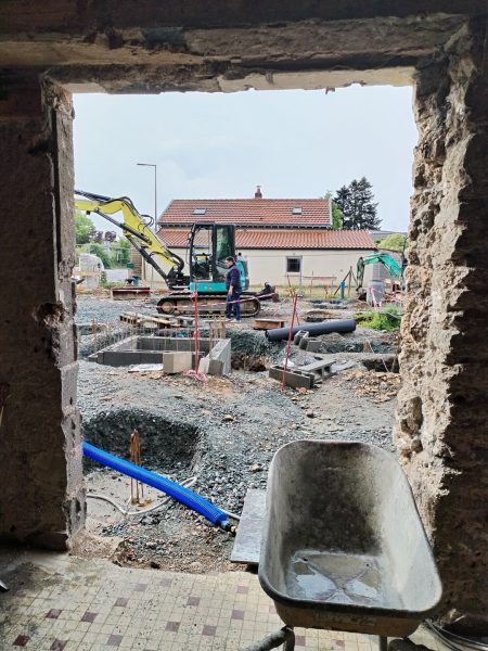 Vue d'un chantier de construction avec excavatrice, fondations en béton et graviers, encadré par une ouverture brute