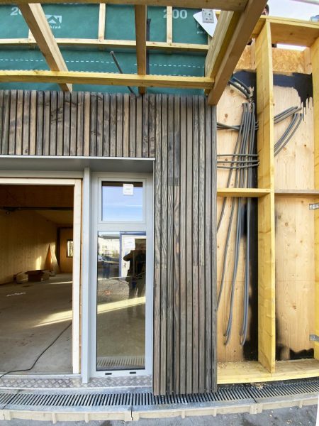 Construction d'une maison à ossature bois avec bardage vertical gris, membrane verte et ouvertures vitrées