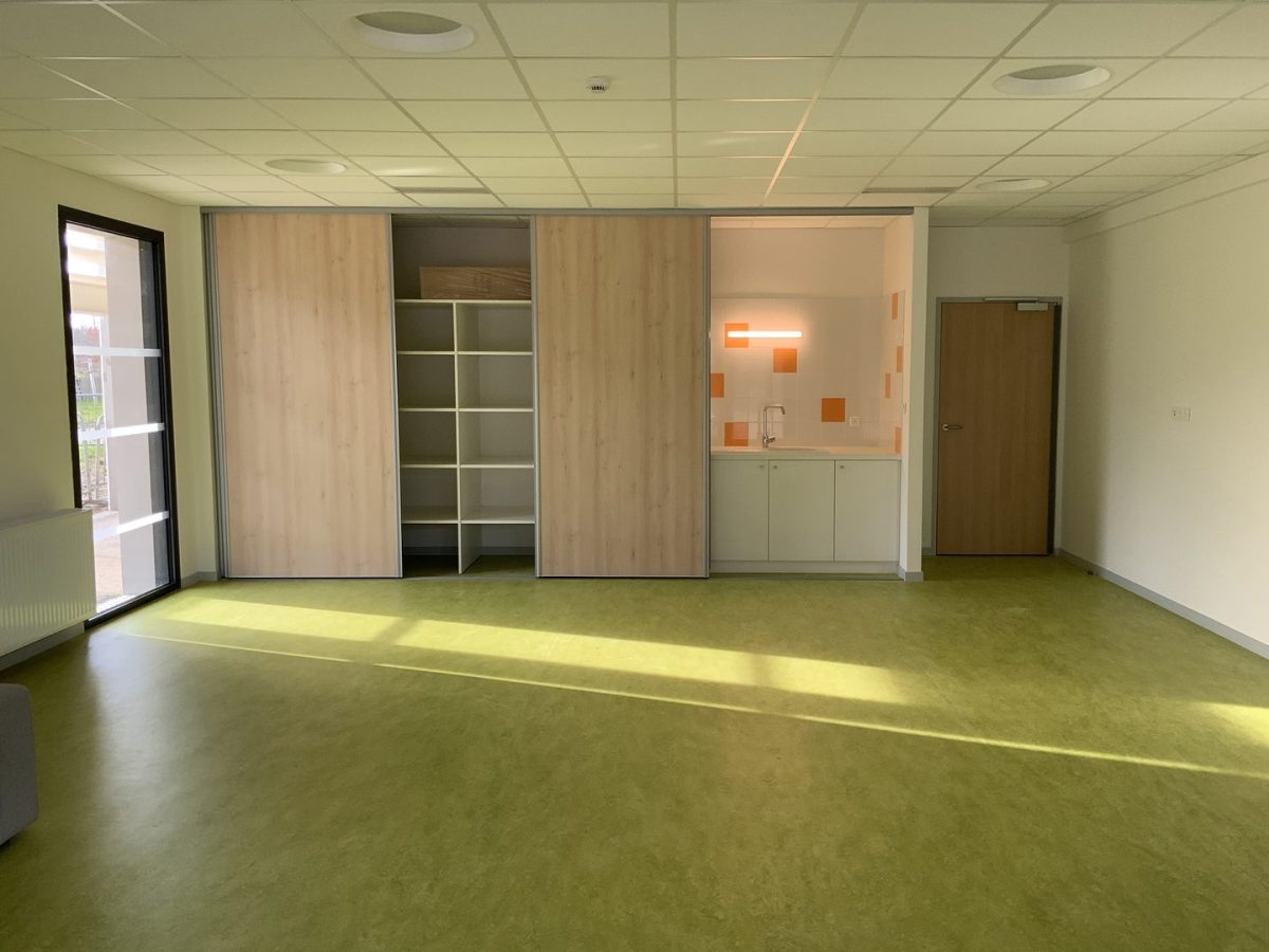 Salle polyvalente avec kitchenette intégrée, rangements coulissants en bois et sol en linoleum vert chartreuse