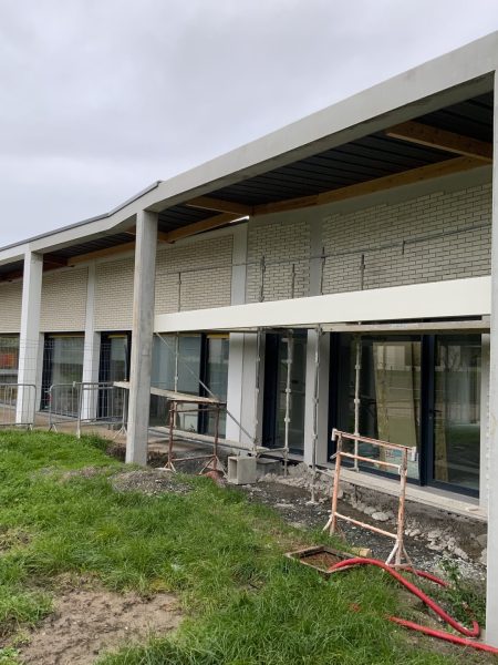 Bâtiment moderne en briques beiges avec toiture plate débordante, grandes baies vitrées et chantier en cours