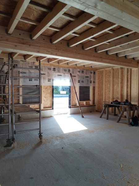 Intérieur en construction avec charpente bois apparente, murs OSB, isolation et baies vitrées donnant sur l'extérieur