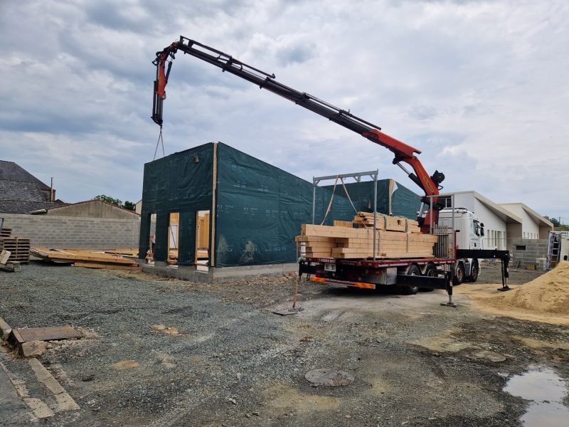Construction d'une maison à ossature bois avec murs enveloppés de bâche verte, grue de chantier livrant des matériaux