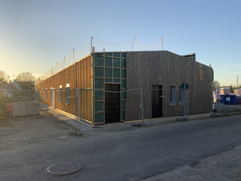 Bâtiment en construction avec façade en bois vertical, conteneurs verts et filet de protection blanc sur le toit