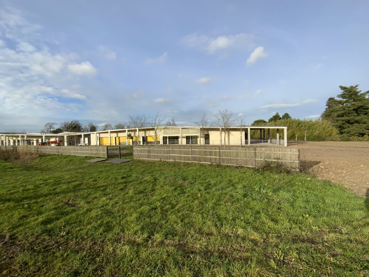 Bâtiment scolaire moderne de plain-pied avec façade beige, clôture bois et espaces verts environnants sous ciel nuageux