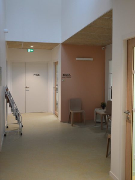 Espace d'accueil infirmier avec mur ocre, faux plafond bois, portes claires et mobilier minimaliste dans un couloir moderne