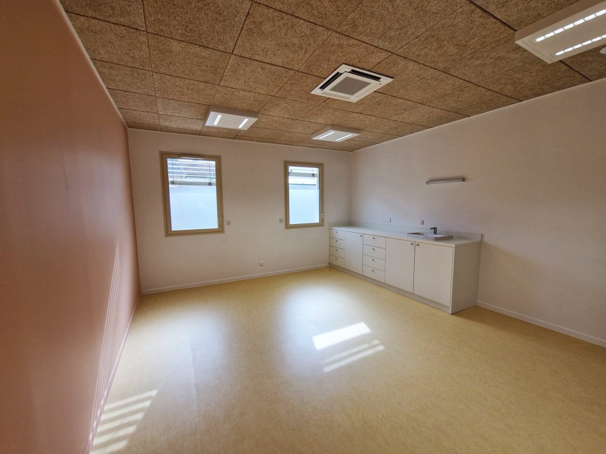 Salle vide avec plafond acoustique, sol jaune, comptoir mural et deux fenêtres à cadre bois