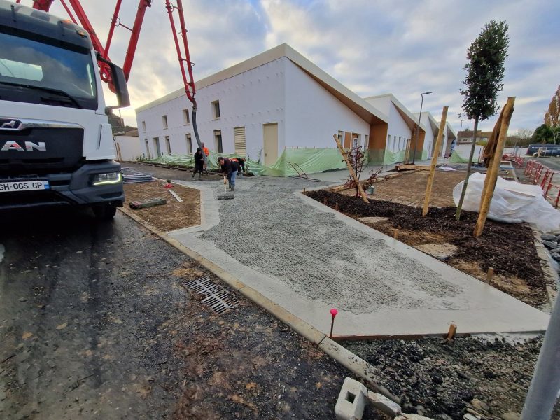 Chantier de construction avec bétonnage d'allée piétonne, bâtiment blanc moderne à toiture double pente en arrière-plan