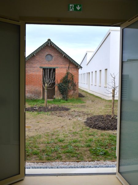 Vue encadrée depuis l'intérieur vers une ancienne grange en brique rouge et un bâtiment moderne blanc avec jardin en cours d'aménagement