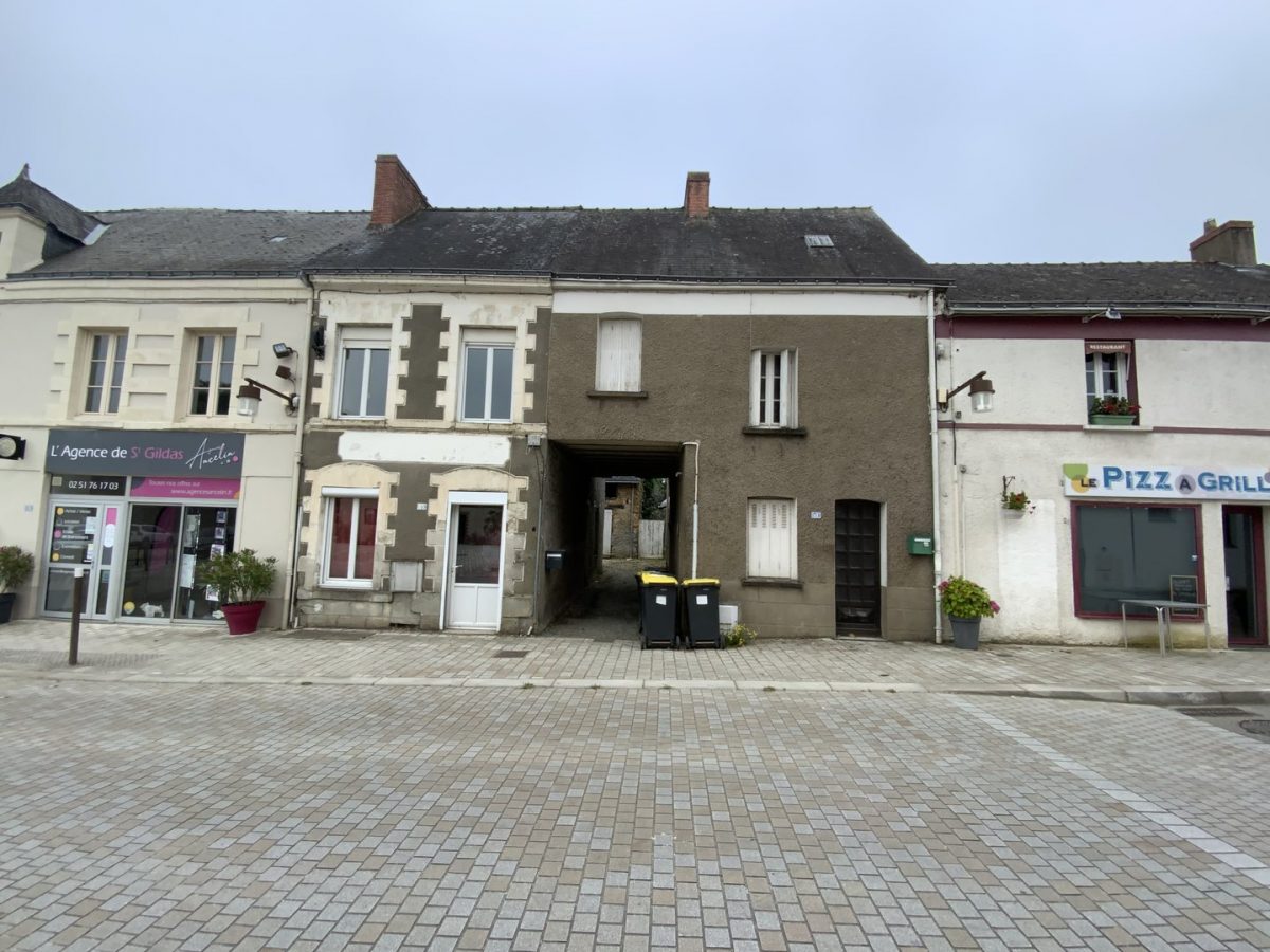 Maisons mitoyennes à deux étages, façades enduites beiges et blanches, toits en ardoise, commerces en rez-de-chaussée, place pavée