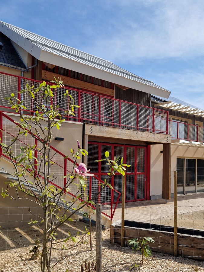 bâtiment enduit avec garde-corps et portails métalliques rouge, au premier plan le jardin avec de la végétation