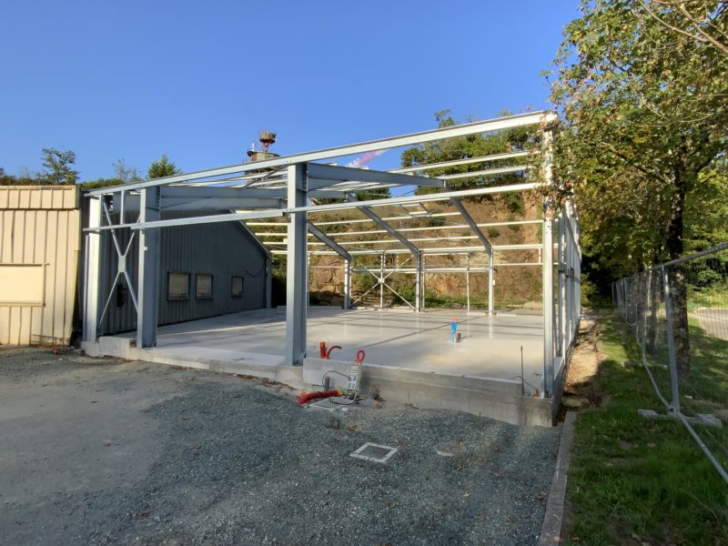 Structure métallique en construction d'un bâtiment industriel, charpente acier apparente, dalle béton, végétation environnante