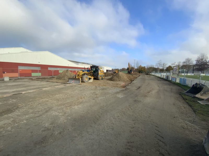 Chantier de terrassement avec engins de chantier près d'un bâtiment rouge et blanc, terrain de sport visible à droite