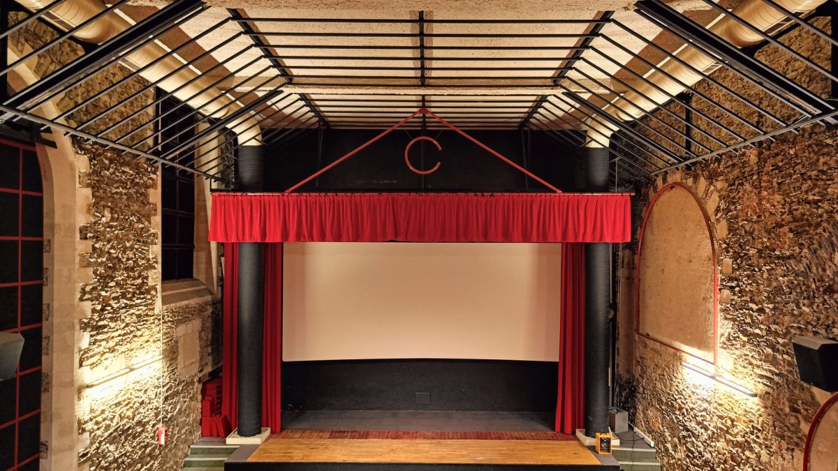 Petit théâtre rénové avec murs en pierre apparente, poutres bois, scène encadrée de rideaux rouges et écran de projection