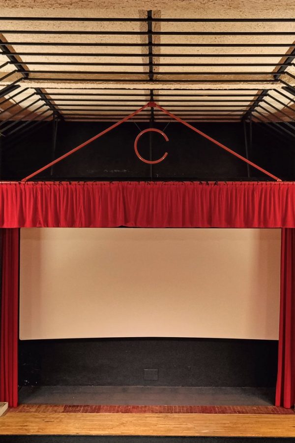 Petit théâtre rénové avec murs en pierre apparente, poutres bois, scène encadrée de rideaux rouges et écran de projection