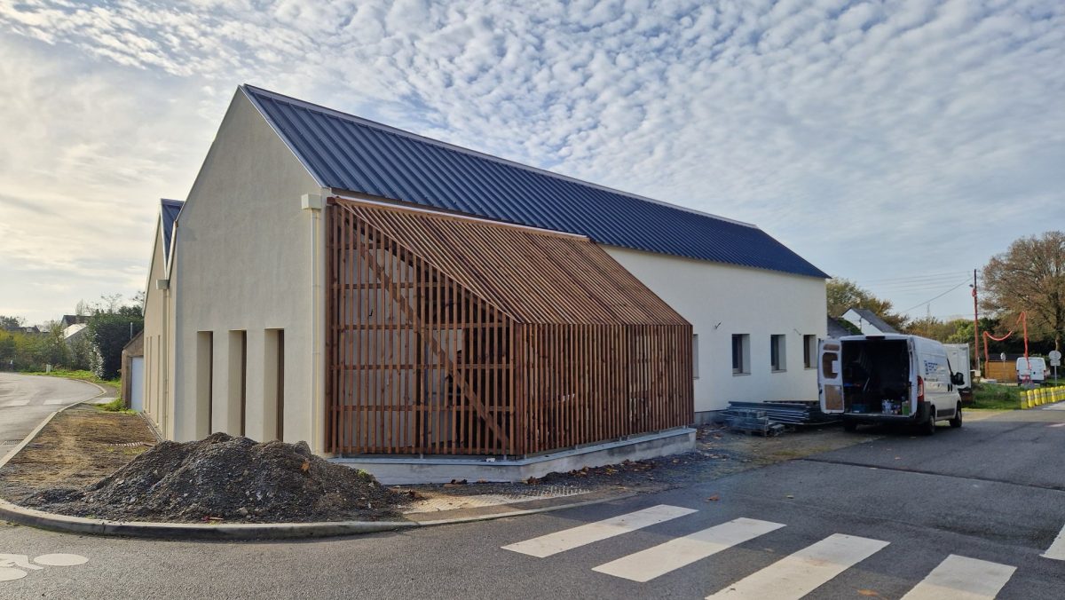 Bâtiment contemporain à pignon avec bardage bois vertical, toiture métallique et enduit blanc, en construction près d'un carrefour