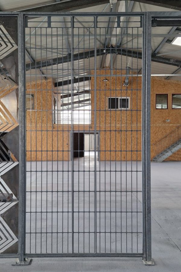 Porte coulissante en acier avec grille verticale ouvrant sur un espace industriel avec murs en OSB et structure métallique