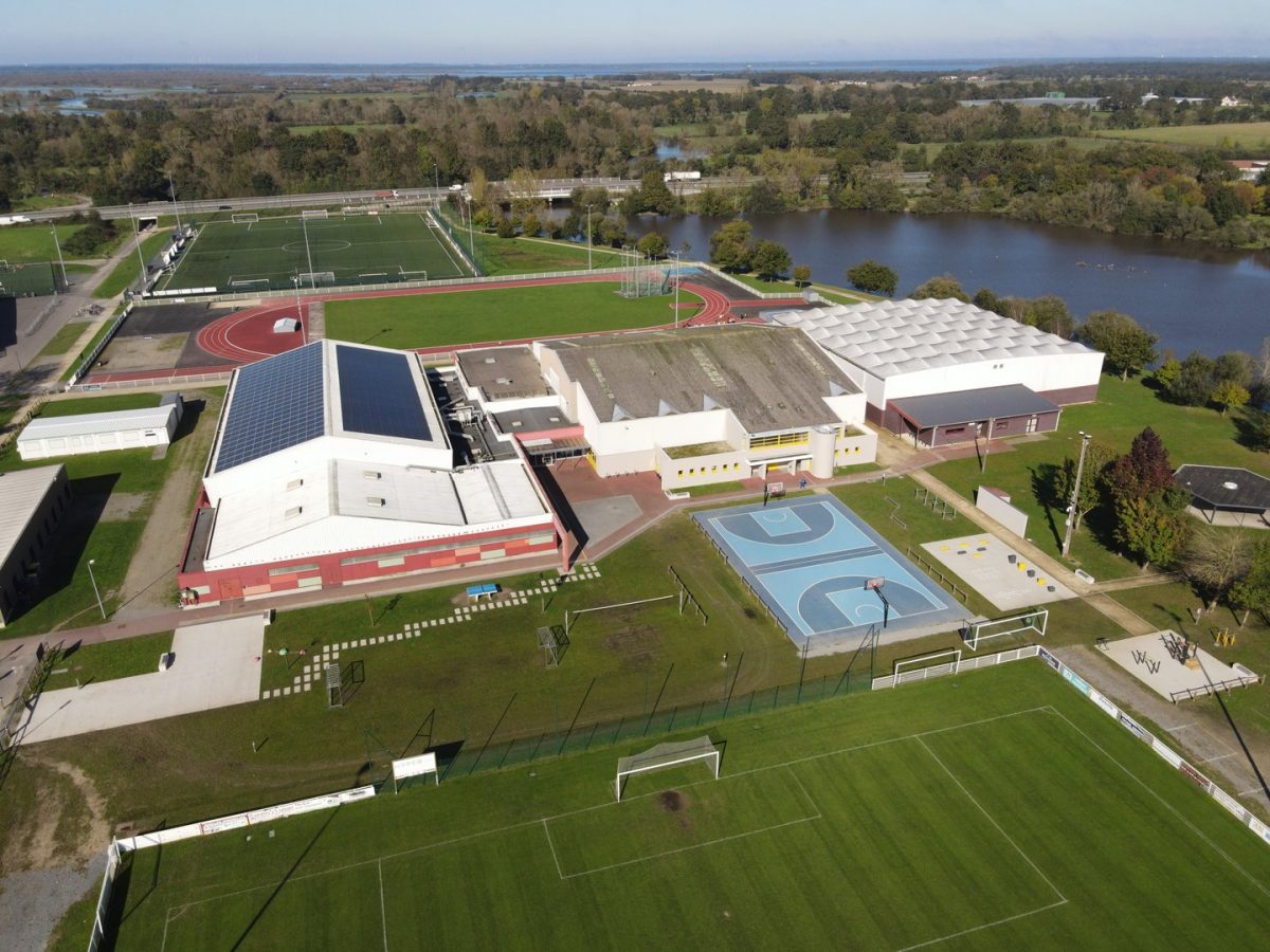 Vue aérienne d'un complexe sportif moderne avec gymnase blanc, terrain de basket extérieur, piste d'athlétisme et lac