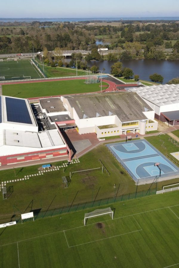 Vue aérienne d'un complexe sportif moderne avec gymnase blanc, terrain de basket extérieur, piste d'athlétisme et lac