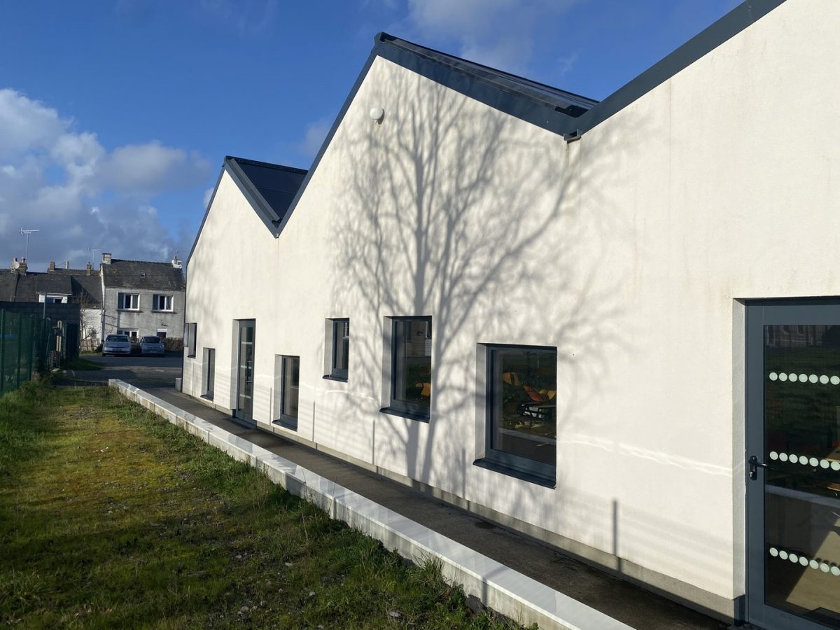 Bâtiment contemporain blanc aux toits sombres avec plusieurs fenêtres rectangulaires et ombres d'arbre projetées sur la façade