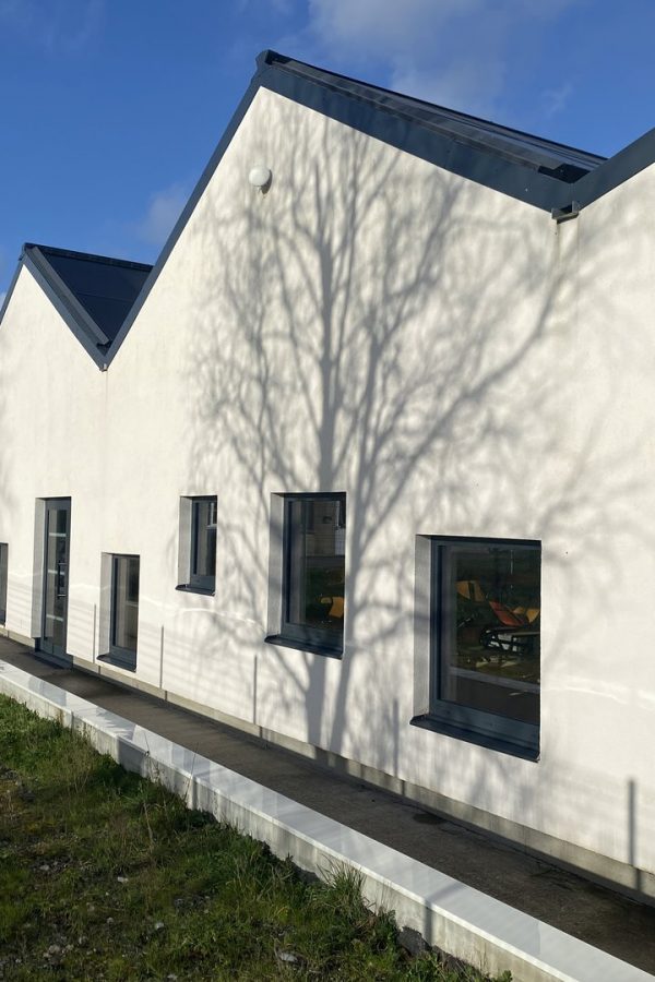 Bâtiment contemporain blanc aux toits sombres avec plusieurs fenêtres rectangulaires et ombres d'arbre projetées sur la façade
