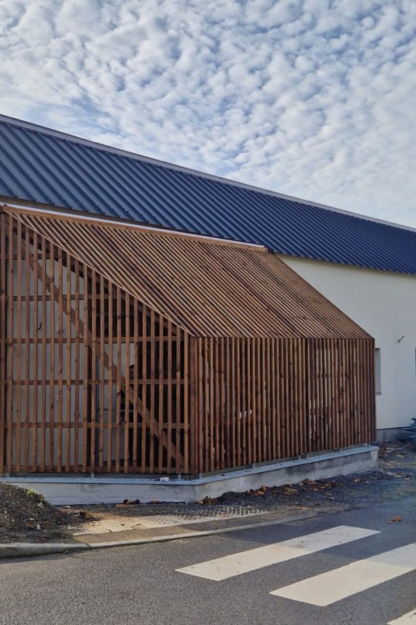 Bâtiment contemporain à pignon avec bardage bois vertical, toiture métallique et enduit blanc, en construction près d'un carrefour