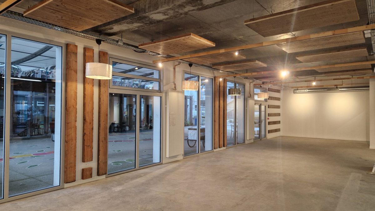 Espace intérieur brut avec plafond béton apparent, poutres bois, baies vitrées donnant sur l'extérieur et sol béton poli