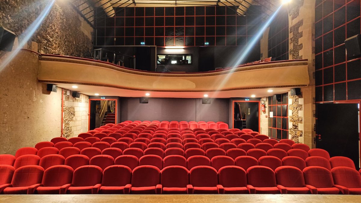 Salle de théâtre avec sièges rouges, balcon bois suspendu, murs pierre apparente et verrière métallique noire