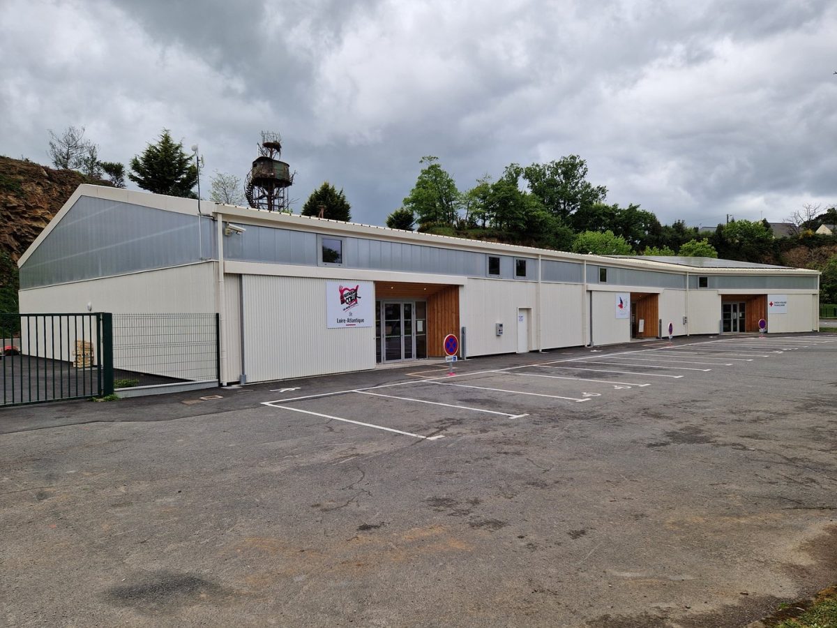 Bâtiment préfabriqué modulaire à bardage métallique blanc avec portes en bois, parking au premier plan, ciel nuageux